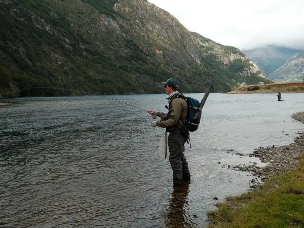 An angler casting where a river leaves a lake in Chilean Patagonia, mountains behind