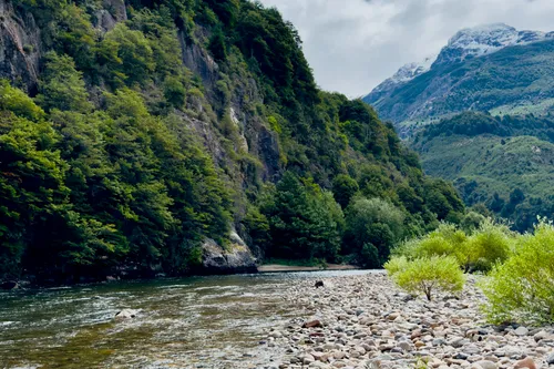 A clear Patagonian river below forested cliffs, a snow-capped peak beyond